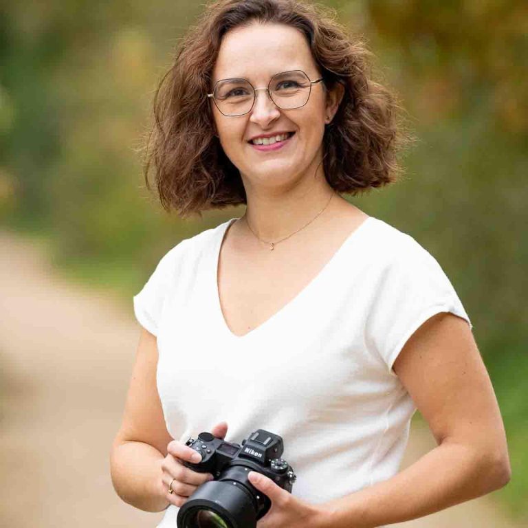 Femme souriante tenant un appareil photo, vêtue d'un haut blanc, en pleine nature.
